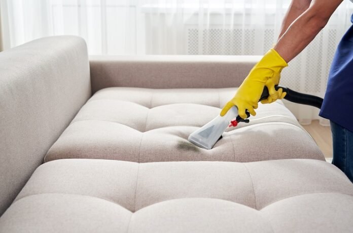 Close-up of housekeeper holding modern washing vacuum cleaner and cleaning dirty sofa with professionally detergent. Professional springclean at home concept